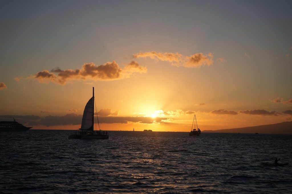 A sailboat and catamaran glide across the Pacific Ocean during a golden sunset, representing a must-do experience to prepare for with your Hawaii packing list.